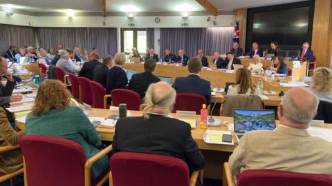 A packed council chamber at Worcestershire County Council - showing several rows of councillors sat on seats, all looking ahead.