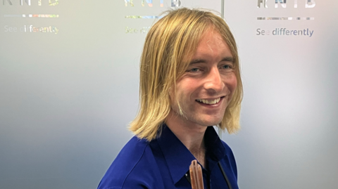 Warren Wilson is standing side on to the camera with his head turned to face it. He is smiling and has medium-length blonde hair. He is wearing a blue shirt. 