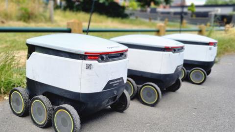 Three small white delivery robots sitting side by side on a pavement.