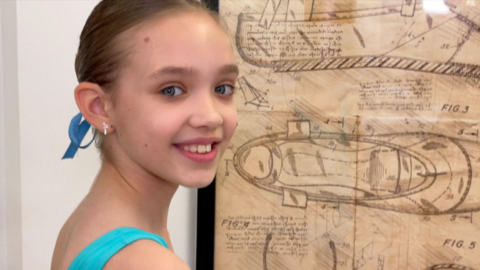Jessica is wearing a blue leotard and is standing in front of a drawn diagram of a ballet shoe, which is stuck on a white wall. She is turn her head over her shoulder, smiling, looking at the camera. 