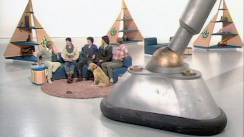 Simon Groom and some of the cast of The Tripods sit on a sofa. A Tripod's leg is in the foreground