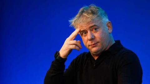 Octopus Energy boss Greg Jackson wearing a black top, against a blue background, with two fingers on his forehead