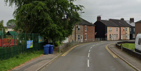 A road, bending to the right, with a children's playground behind a fence on the left of the road, followed by terraced houses