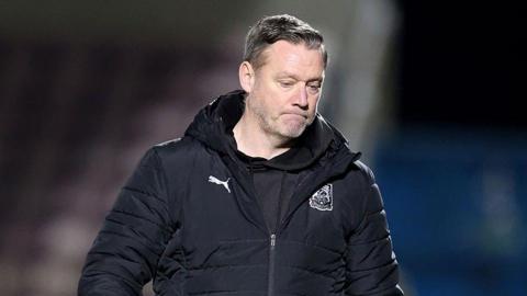 A man in a black sports jacket frowns whilst looking at the floor.
