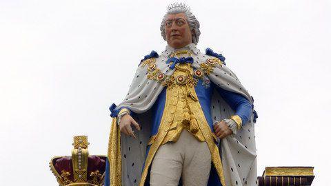 A close-up of the King George III statue. The statue is painted in lifelike colours, with rosy cheeks and eyebrows. The king is wearing a spotted ermine robe, an ornate gold chain of office, a blue and gold coat and white briches. His right hand is empty but the thumb and fingers are curled around as though they should be holding something.