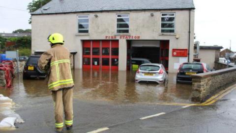 A firefighter in uniform stands in front of a fire station building - a beige stone building with fire station signage. The building's car park is flooded with brown water and several cars are partially submerged in water.