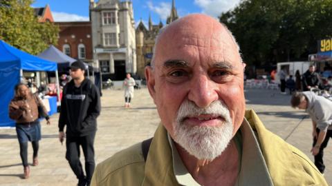 Toby Wood smiling into the camera while standing in Cathedral Square, with people walking behind him. He has a bald head and a white beard and moustache, and is wearing a beige jacket.