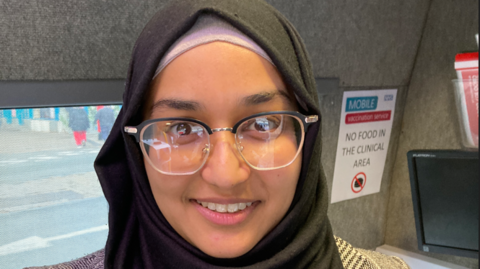 A woman in a black headscarf with large glasses smiles towards the camera. Behind her is a grey wall with a poster that says no food in the clinical area