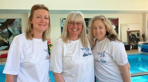 Three women stood shoulder to shoulder by a pool wearing white t shirts.