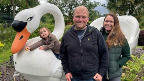 A man, woman and child in a garden in front of a large white boat in the shape of a swan, with orange beak and black nose.