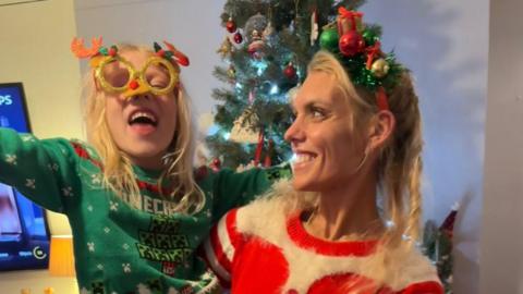 A woman in a red and white Christmas jumper and green Christmas hairband, carrying a young child. The girl is wearing a green Minecraft Christmas jumper and novelty glasses. They are pictured in front of a Christmas tree.