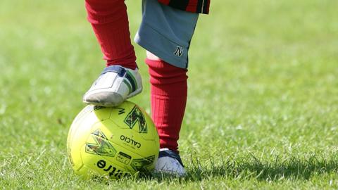 A child is balancing a yellow football on the grass with his foot on top of it. You cannot see the child's face. They are wearing red socks and white trainers 
