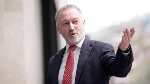 Steve Reed, a man in his late fifties with short grey hair and a white beard, speaking and looking to the right wearing a suit and red tie pointing his left arm towards the camera.