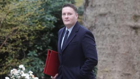 Streeting walking outdoors in what appears to be a landscaped garden or park area. He is wearing a long, dark overcoat and holding a bright red folder under one arm. The background features dense greenery and flowers.