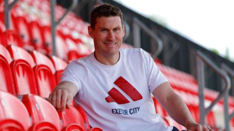 Matt Taylor is wearing a white T-shirt with Exeter City on the front and sitting in a stadium.