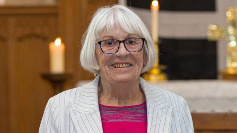 Brenda Wright has short grey hair and is wearing a pair of glasses, a white jacket, and a pink top. She is smiling.