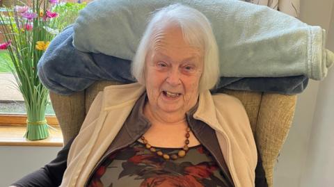 A woman with white hair and a beaded necklace with a gillet on that is light brown cream, looks at the camera from an armchair, with a vase of flowers on the window sill behind her. 