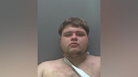 Police custody shot of Levi Baxter looking at the camera. He has short mid-brown hair. He is topless and appears to have a sling around his neck