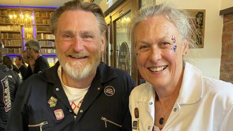 A man and a woman smiling at the camera. He is to the left and has brown hair and a grey beard, she has grey hair tied back. They are both wearing boiler suits - him navy, her white. She has red blue and silver glitter on her face, to the outer side of her eyes.