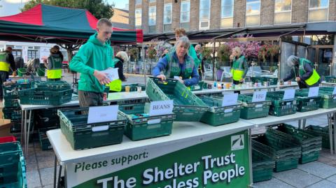 There's a row of white tables with green crates on them with labels on them showing what types of tins should be placed in the crates like beans and veg. A man and a woman are sorting out some of the creates at the front while more volunteers work in the background.