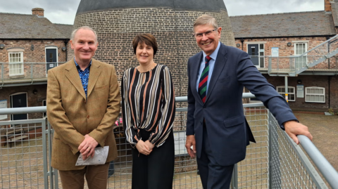 A woman is standing outdoors in between two men. They are positioned against a set of metal railings and looking towards the camera. Behind the group is a bottle-shaped bottle kiln made of brick and a series of two-storey factory buildings.