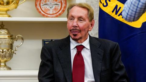 Oracle co-founder Larry Ellison sitting in a chair at the White House wearing a dark grey suit, white dress shirt and deep red neck tie.
