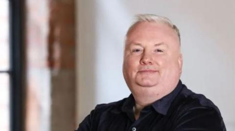 Stephen Nolan filmed in a room with red brick and white walls. He has short grey hair and a black shirt. He is standing with folded arms