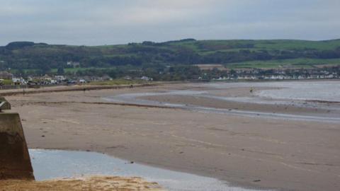 Ayr beach - a large expanse of sand, a grey sea. Green hill in the background and grey sky.