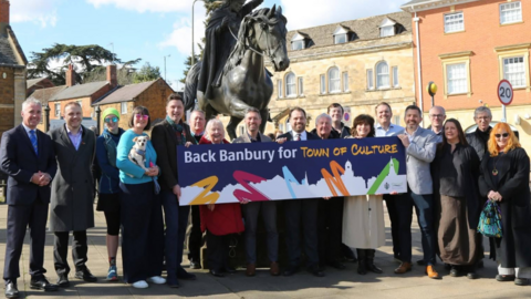 A line of people stood in front of a statue of a lady riding a horse, with some holding a large sign saying 'Back Banbury for Town of Culture'