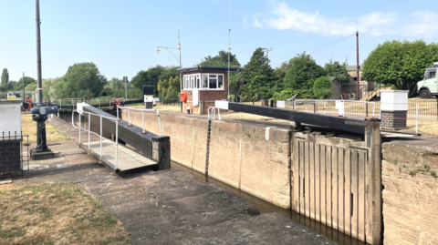 The lock is pictured open and against the side of the river. A brick wall is on the opposite side in the river and a small building with windows stands also on the right side of the water.