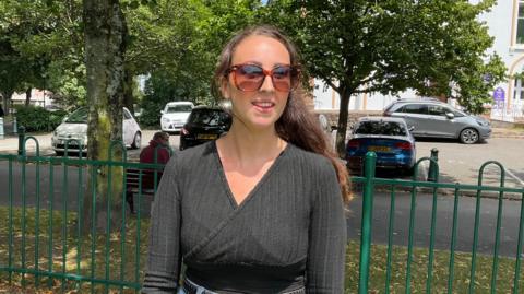 Sarah Aubert - A woman with long brunette hair, she's wearing big brown sunglasses and a black top. 