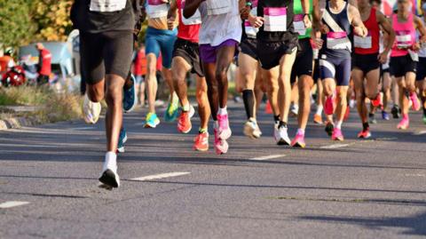 A photo from ground level of a group of runners' legs running on the concrete road. They are wearing shorts with colourful shoes.