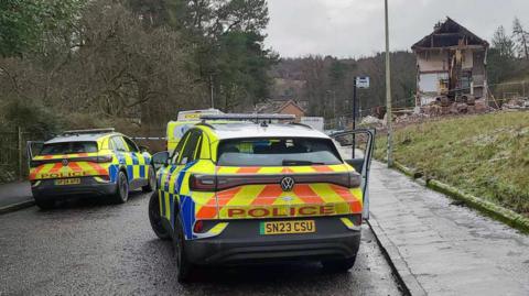 Two police vehicles parked on a residential road near a severely damaged house, with debris visible and trees lining the street