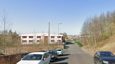 A street view, showing a large warehouse building with a road lined with cars and a grassy bank with trees on either side.