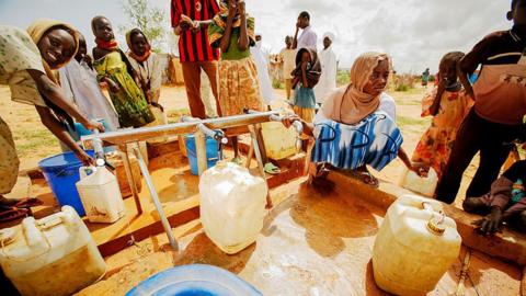 People in a desert landscape pouring water from pipes into plastic jugs.