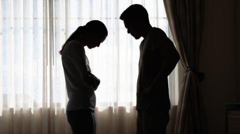 The silhouettes of a man and a woman standing in front of a window with sheer curtains drawn. The woman is holding her arms around her waist and looking down, while the man has his hands on his hips and appears to be shouting at her.