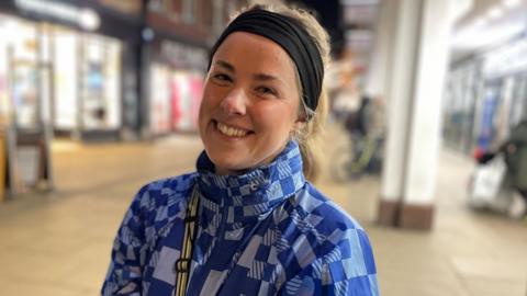 Laura-May Land wears a black hairband, a blue patterned jacket and has a strap across her shoulder. She is smiling directly at the camera and has her head tilted to the side. She is standing in Cambridge city centre.