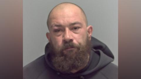 A custody image of a man with a shaved head and a thick beard. He is wearing a grey hoodie.