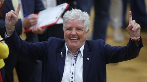 Alison Bennington smiling and raising her index fingers above her head 
while celebrating winning her seat in the 2023 council elections.  She is wearing a navy blazer and a white shirt with blue buttons.