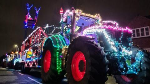 A tractor is lit up in many different colours for Christmas, with fairy lights around its wheels and on a trailer and over the front of the vehicle.