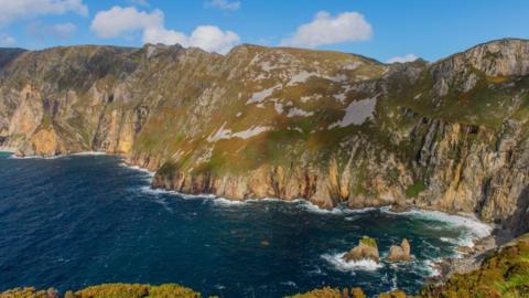 Slieve League cliffs. Beneath them a sea with some white water around the cliffs and above them a blue sky with some clouds.