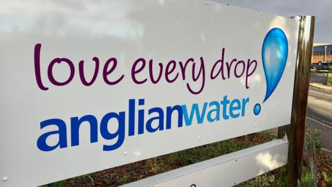 A white sign with purple and blue writing. It reads 'Anglian Water, Love every drop'. There is a exclamation mark at the end which had been designed to look like two droplets of water. The sign is outside the companies HQ