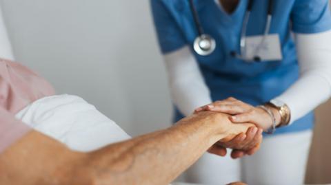 A close-up image of a care worker holding a patient's arm while they lie in bed.