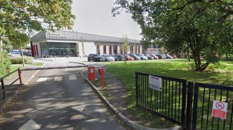 A road with trees on either side is leading to a large building with a number of cars parked in front. 