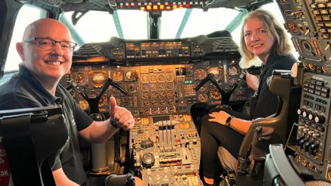 Carolynne and Richard Hutchins sitting in the cockpit of the supersonic aircraft with an array of control panels and switches around them. The couple are smiling and Richard is doing a thumbs up.