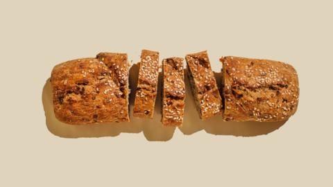 Loaf of wholemeal and seeded bread. Three slices in the middle have been cut. The bread has a shadow coming off it and it's on a pale beige background. 