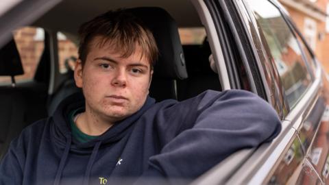 Young 18 year old man sits in the passenger seat of a black VW car, his arm resting on the open window as he looks to camera