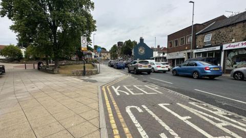 A road with two lanes of cars travelling on it. An area of the road is marked 'Keep Clear'. A paved pedestrian area on the left side with a small patch of grass and a tree. A row of shops on the opposite side of the road.