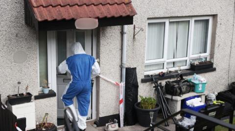 An individual in a forensic protective suit is walking into a property