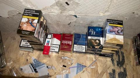 A pile of different brands of packets of cigarettes piled up against a damaged white wall and on a wooden floor.